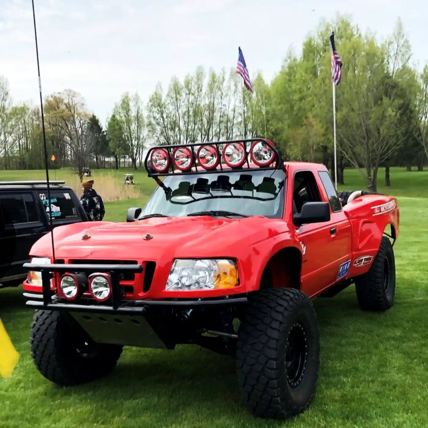 Custom red off-road truck with oversized tires, roof-mounted rally lights, and STEELE Racing Products branding, parked on a grassy field with American flags in the background